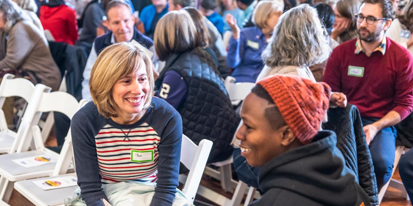 Photograph of a large room filled with people sitting in white folding chairs. In the foreground, two people smiling are seen in conversation.
