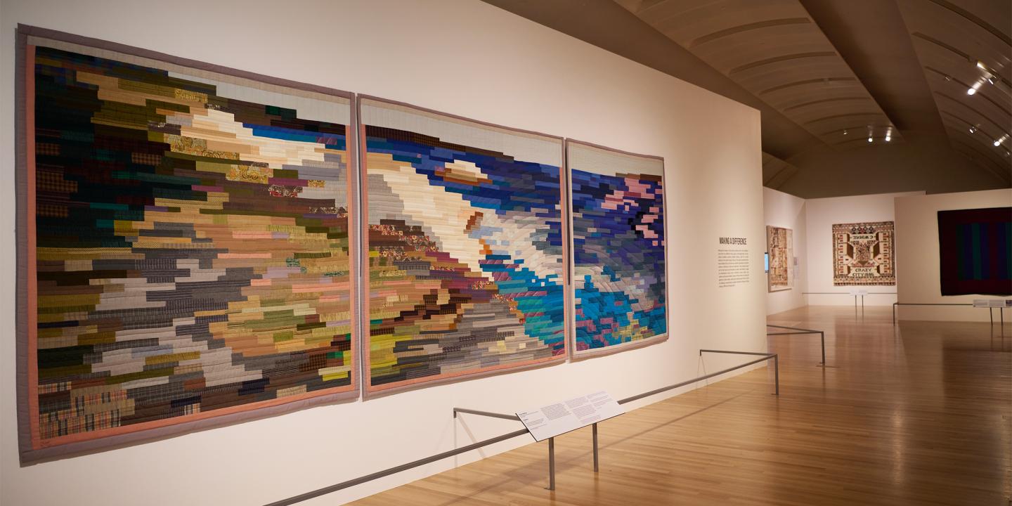 Image of a gallery space. Colorful quilts are seen hanging on white walls. Closest to the frame is a triptych quilt displaying a coastline and ocean made up of colorful bands of fabric.