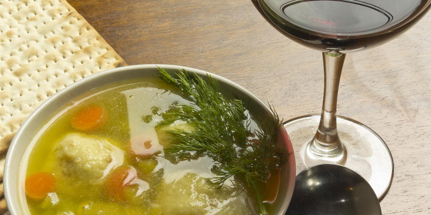 Overhead photo of a bowl filled with matzo ball soup. Next to the bowl is an unleavened cracker, glass of red wine, and spoon.