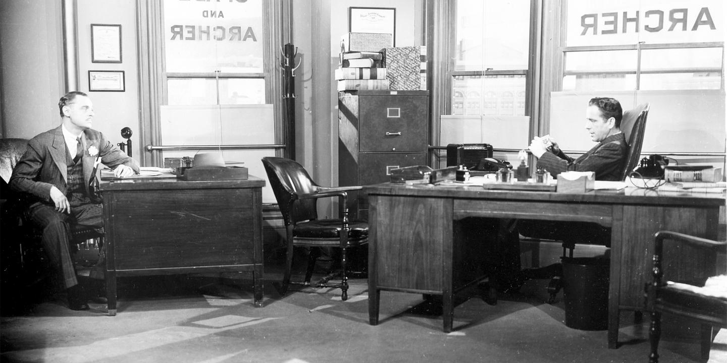 Black and white image of two men sitting at desks in an office conversing. The name "Archer" is painted on the windows behind them.