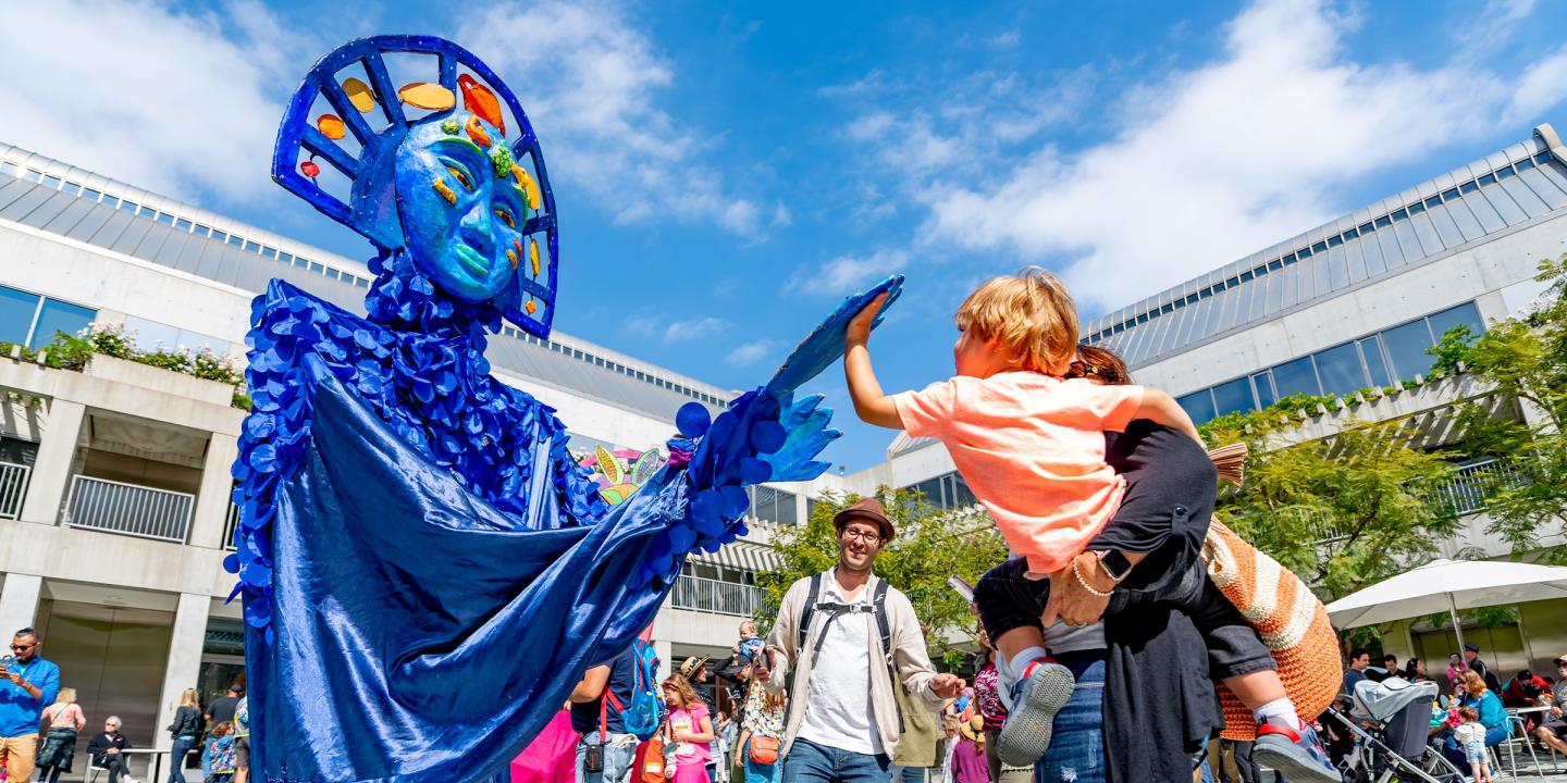 Outdoor photo at the Skirball campus. A large, blue puppet is on the left, reaching out to a young child in their adult's arms and is reaching back. People can be seen behind the trio dancing.
