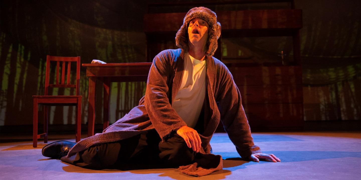 Photograph of a man kneeling on a stage looking up. He is dressed in a long, brown robe and wearing a hat that looks like floppy dog ears.