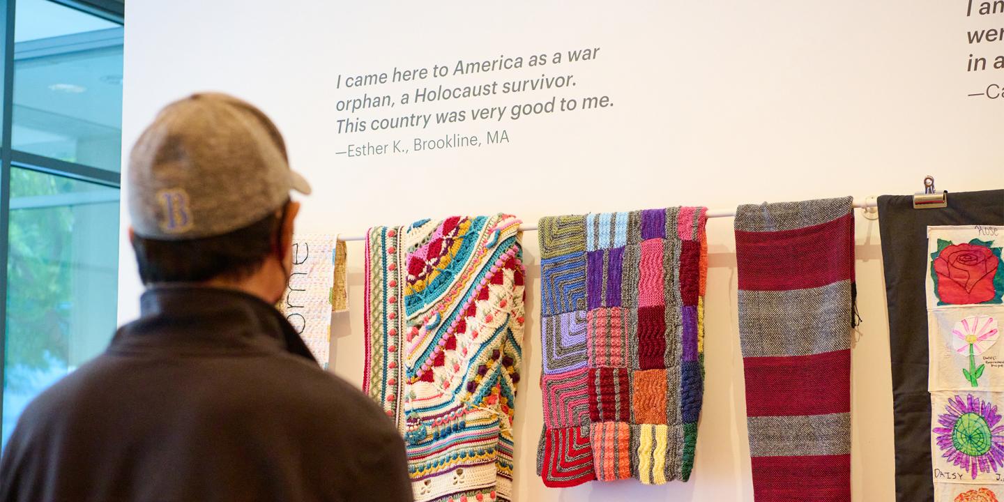 Photo of a man in a baseball cap facing a white wall with many colorful blankets displayed.