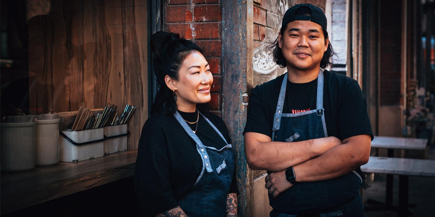 Two people standing in front of a brick building with cutlery out in pails. They are wearing black shirts and blue aprons smiling.