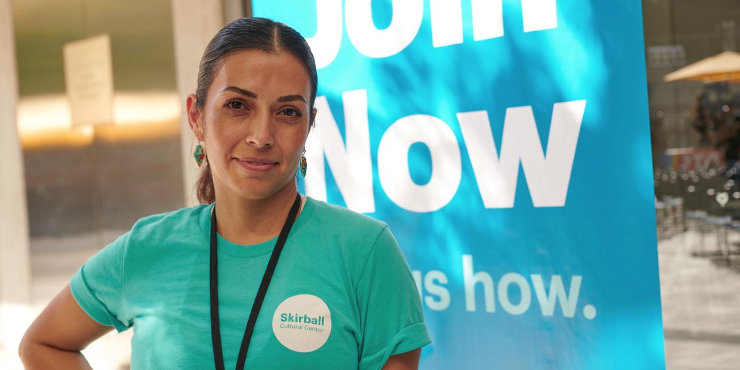 Skirball employee with Skirball branded teal t-shirt standing and smiling in front of a Join Now Skirball banner.