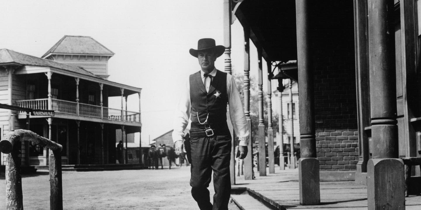 Still from the movie showing Gary Cooper walking towards the camera wearing dark clothing and a cowboy hat. He is surrounded by western buildings and a bright sky.