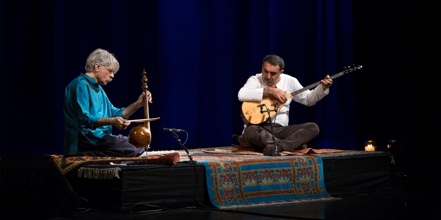 Photo of a dark stage. Kayhan Kalhor and Erdal Erzincan sit on a rug with instruments playing together.
