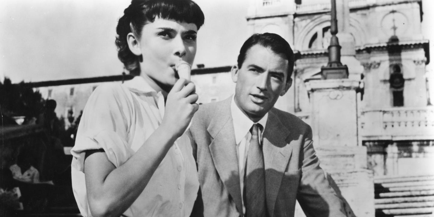 Still from the film Roman Holiday showing Audrey Hepburn and Gregory Peck outside on a street in Rome. Hepburn is eating ice cream, and Peck is looking at her.