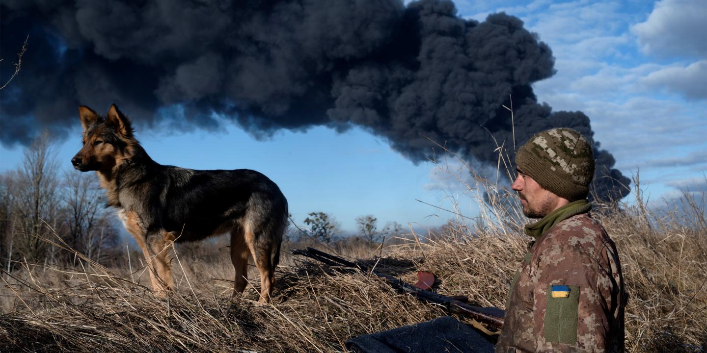 Photo of a Ukrainian soldier in a field with his dog. Both are looking at something off camera to the left. Behind the man is a large plume of black smoke filling the skyline.