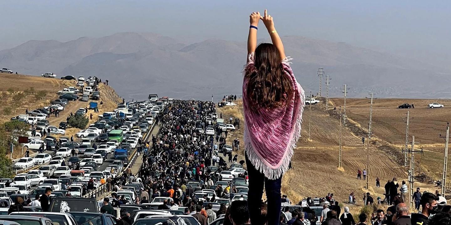 Woman standing on a car with arms up high, looking a the line of stopped cars that goes beyond the horizon. The landscape of Iran beyond.