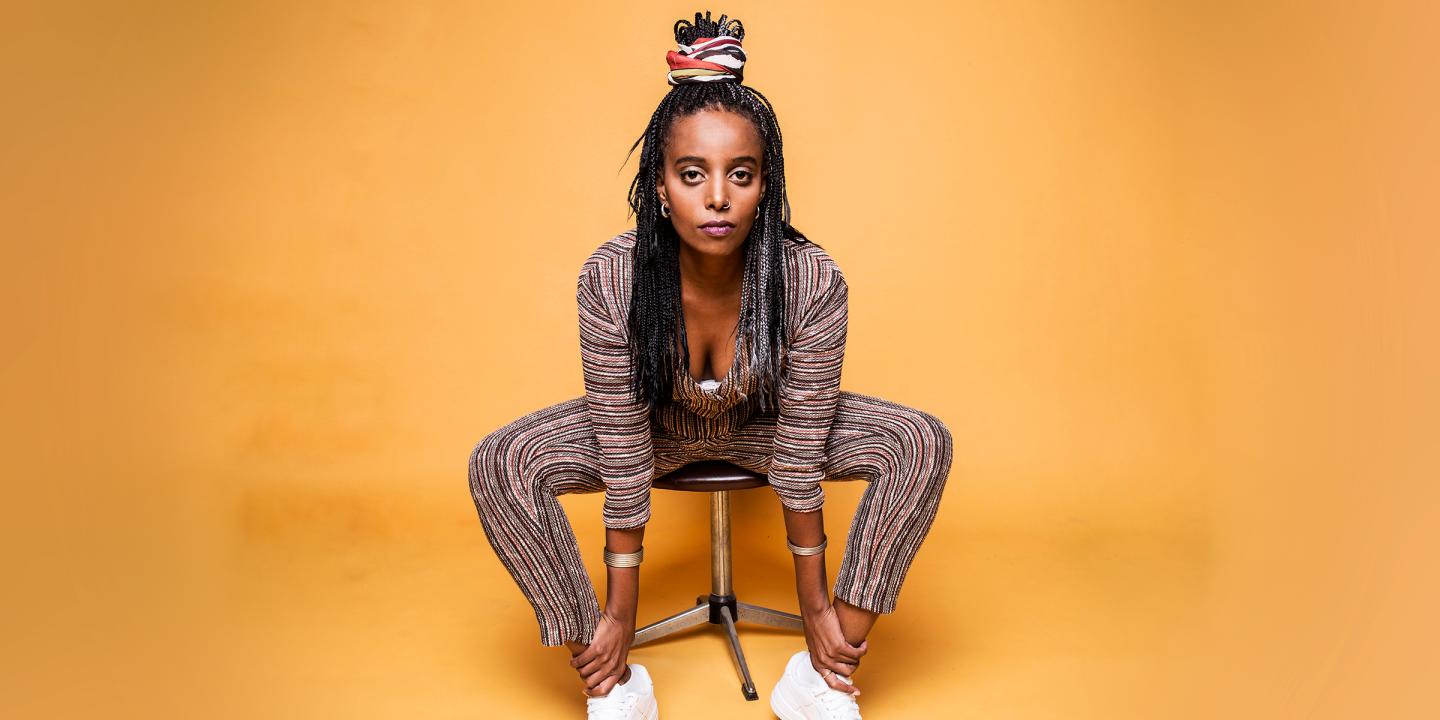 The artist sitting on a stool in front of a yellow backdrop. They are looking directly to camera.