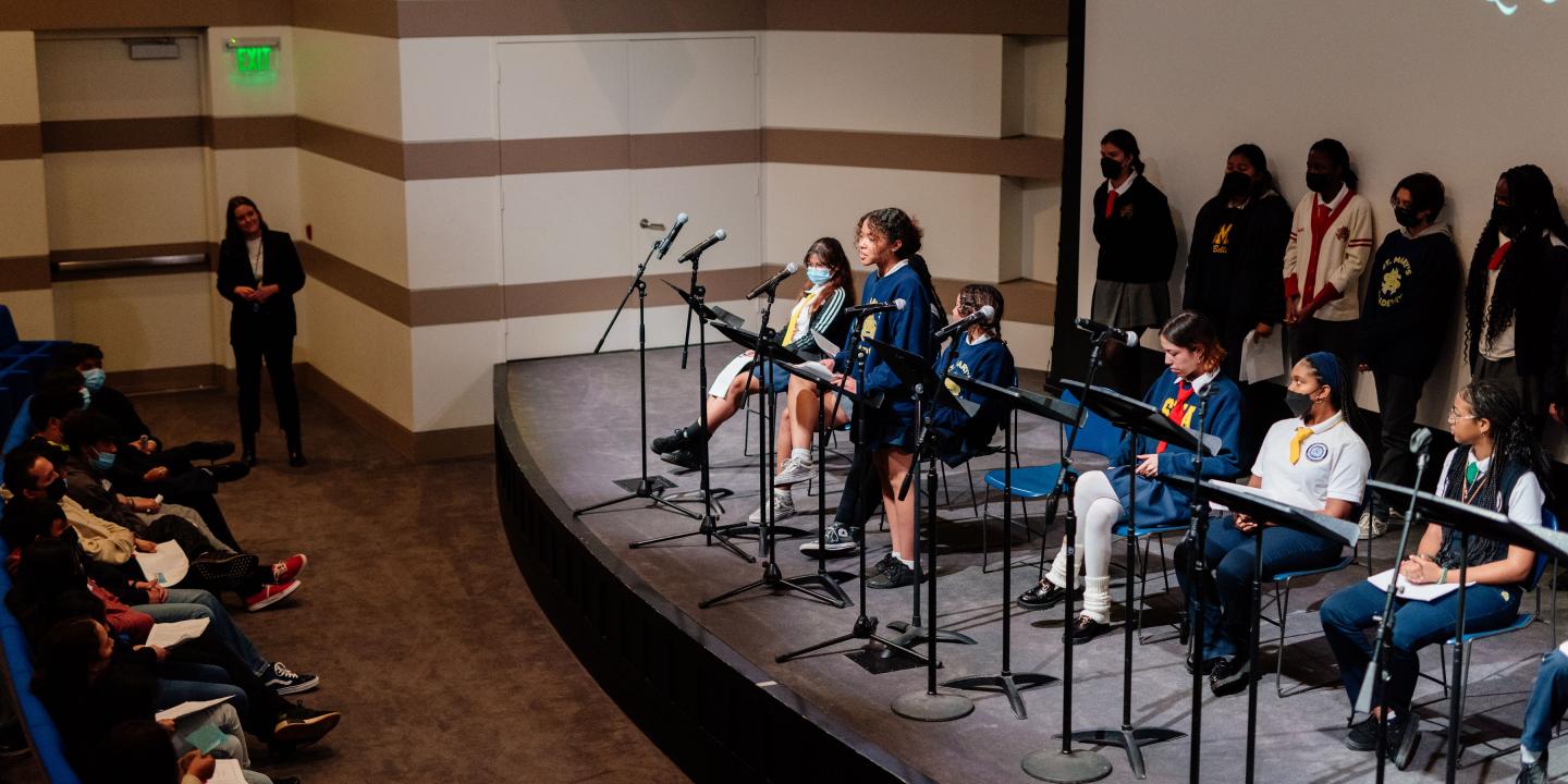 Photo of a group of students lined up on a stage. One student is standing in front of a microphone speaking to the audience.