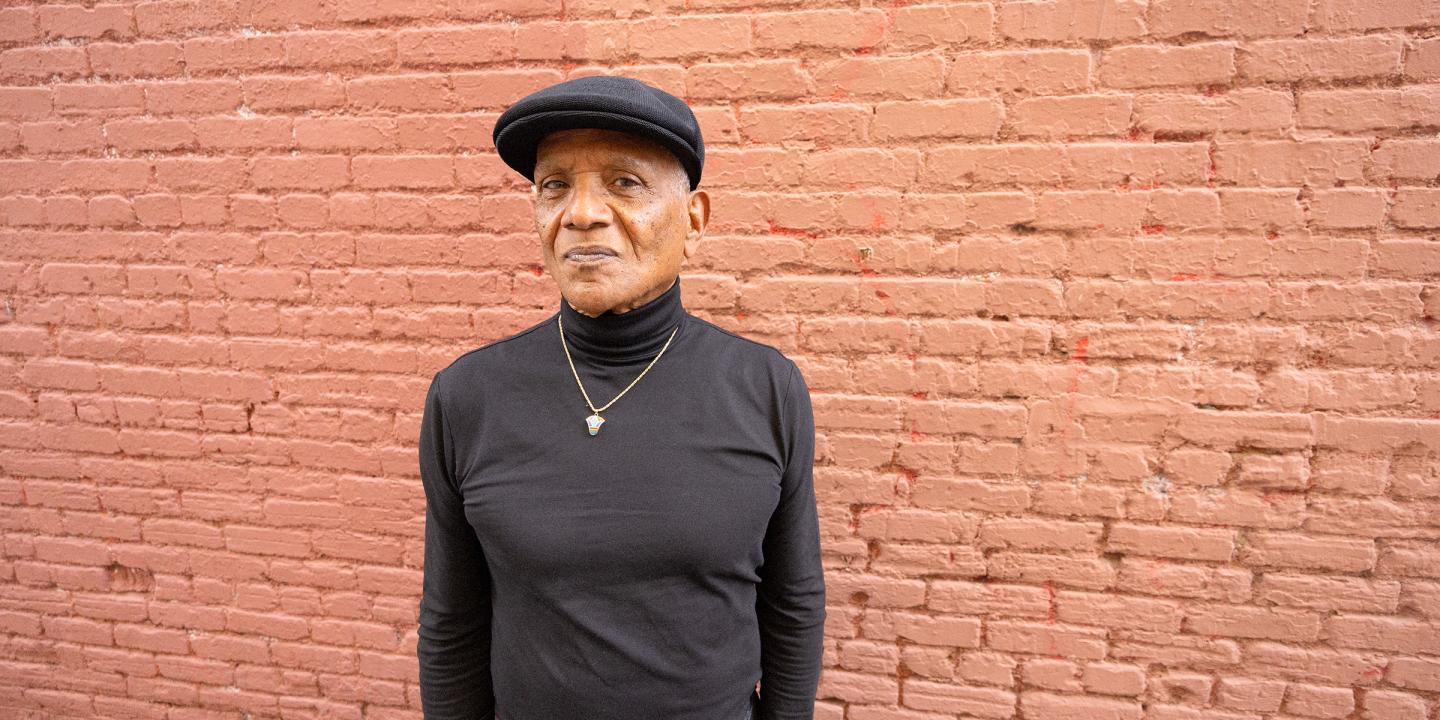 The artist dressed in all black and standing in front of a red brick wall looking off camera.