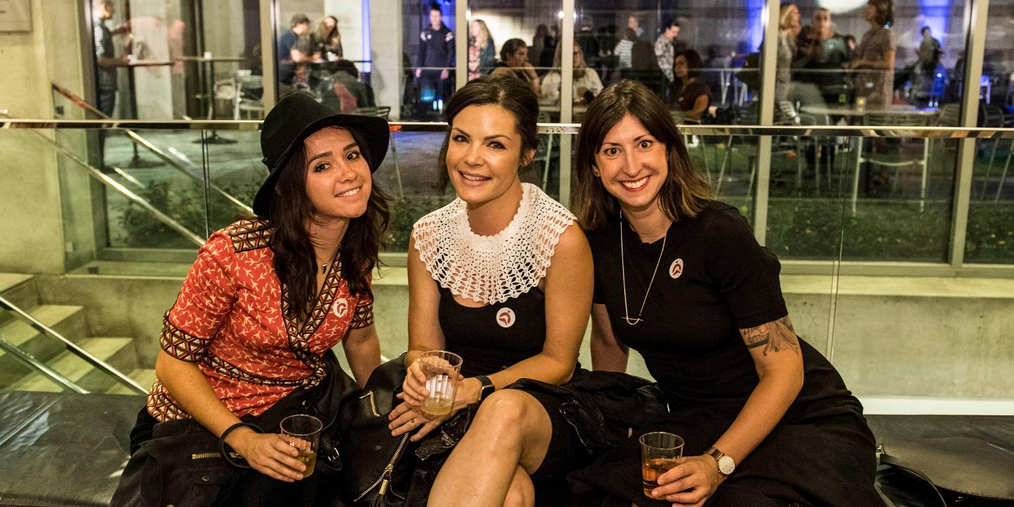 Three women sit together on a bench smiling at the camera. Behind them a party can be seen through a wall of windows.