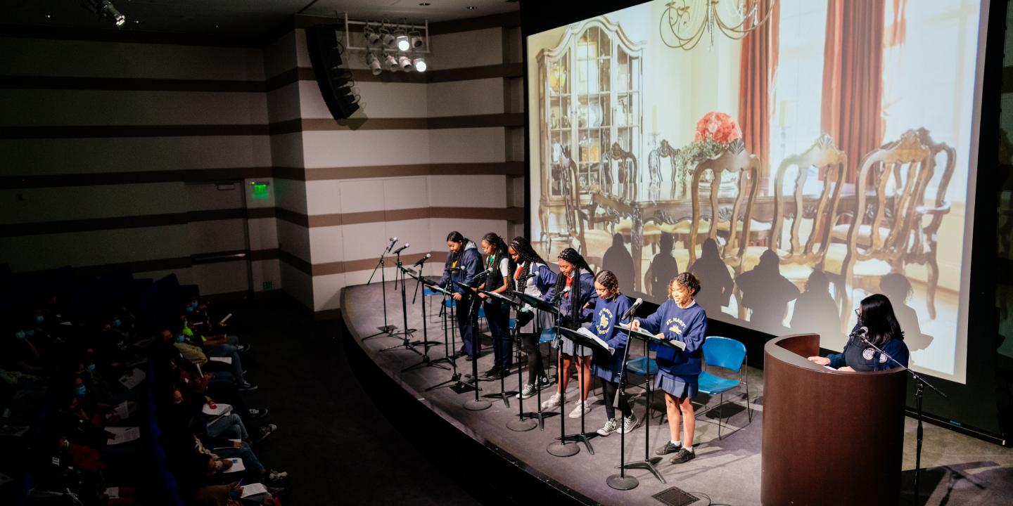 Photo of a group of students lined up on a stage with microphones and stands presenting to a room of people.