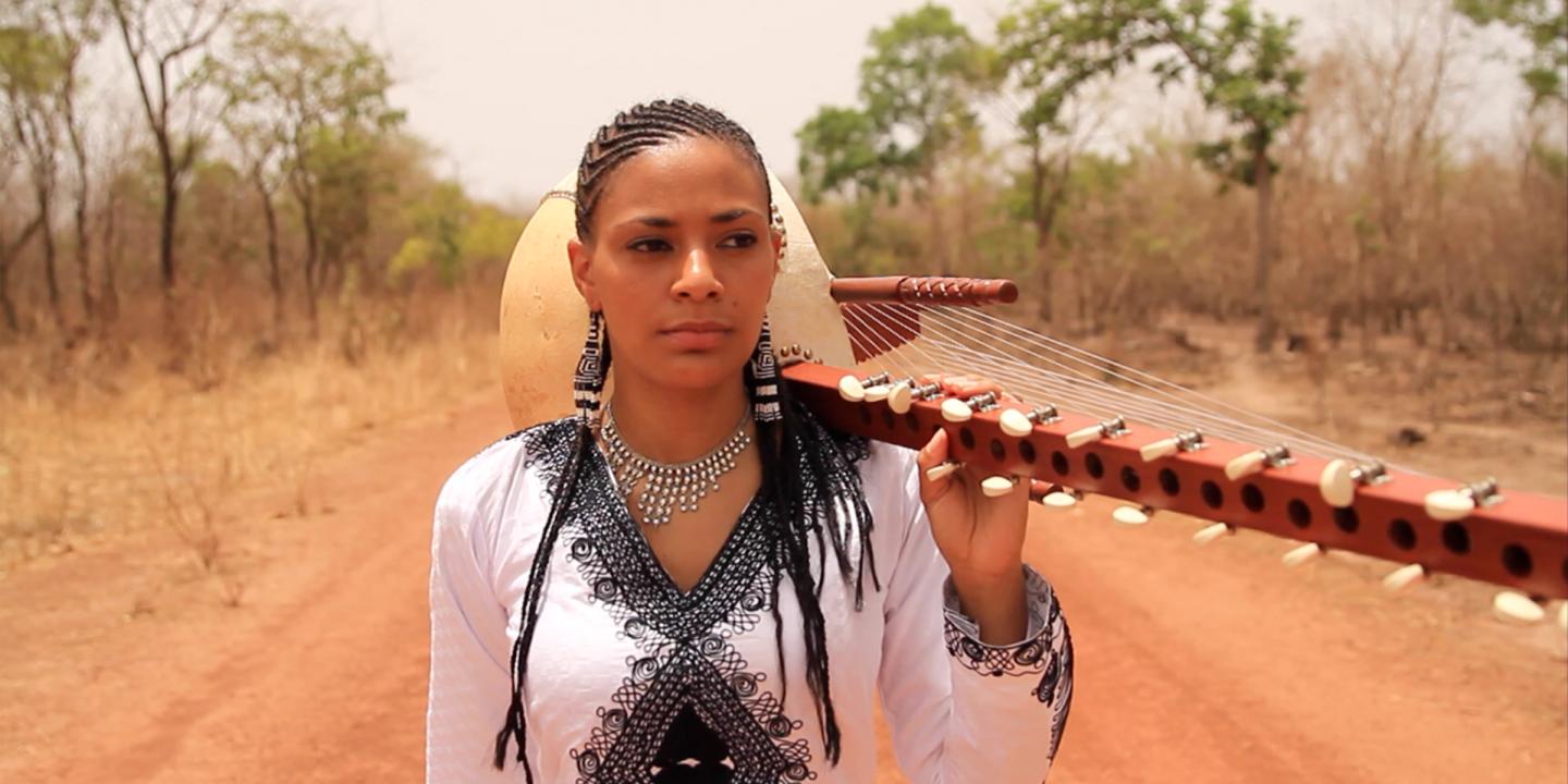 Colorful photograph of Sona Jobarteh standing on a dirt road lined with trees. She is holding her kora over her shoulder and looking slightly away from the camera.