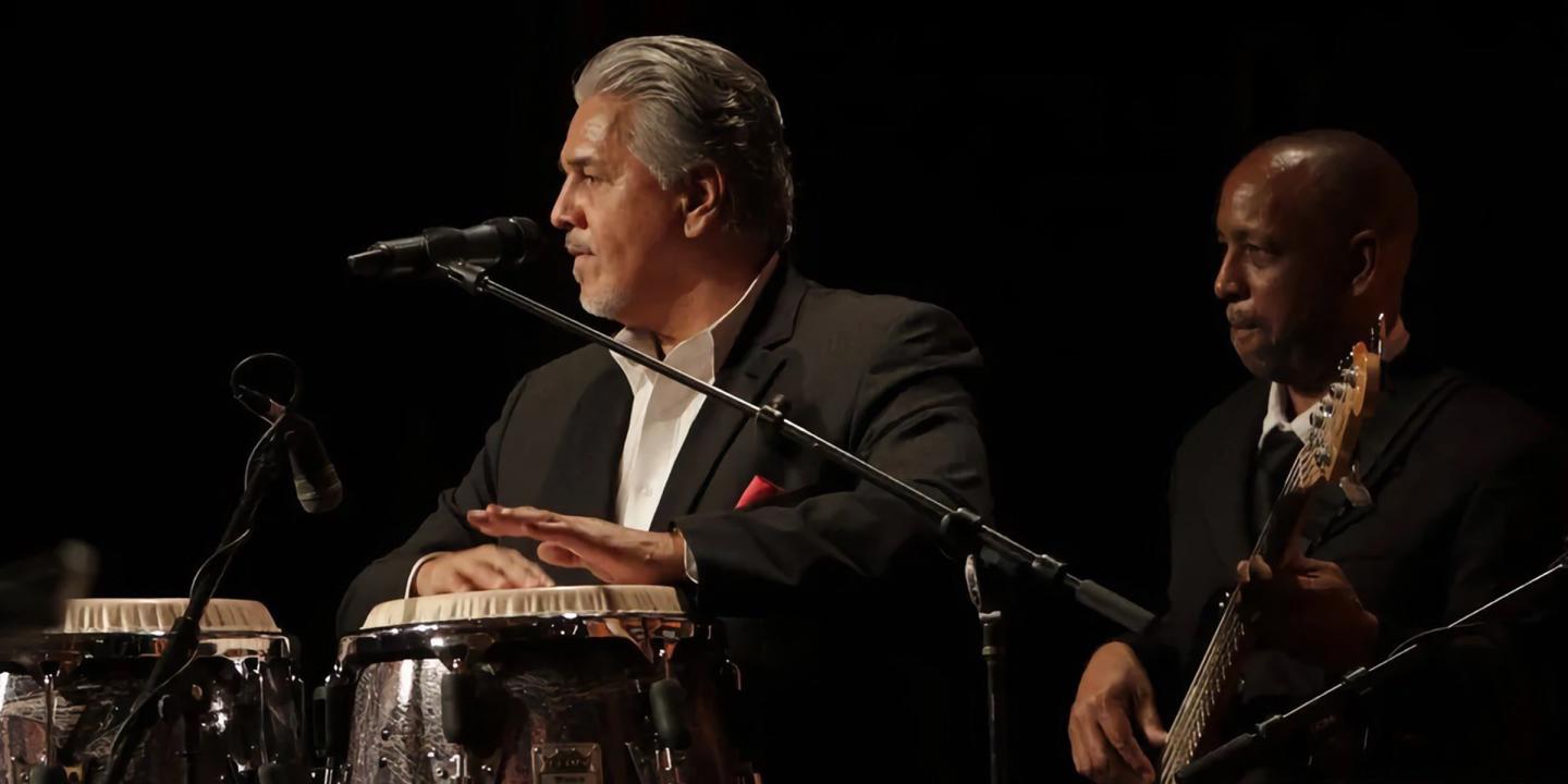 Photo of the artist on a dark stage playing his instrument with another musician behind him.