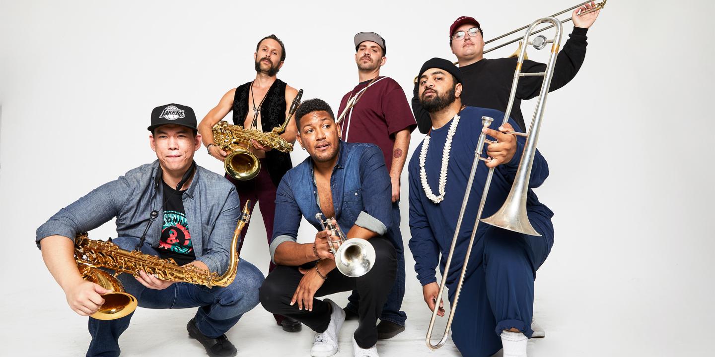 Photo of the six band members standing and kneeling in front of a white background. Each member is holding their instrument.