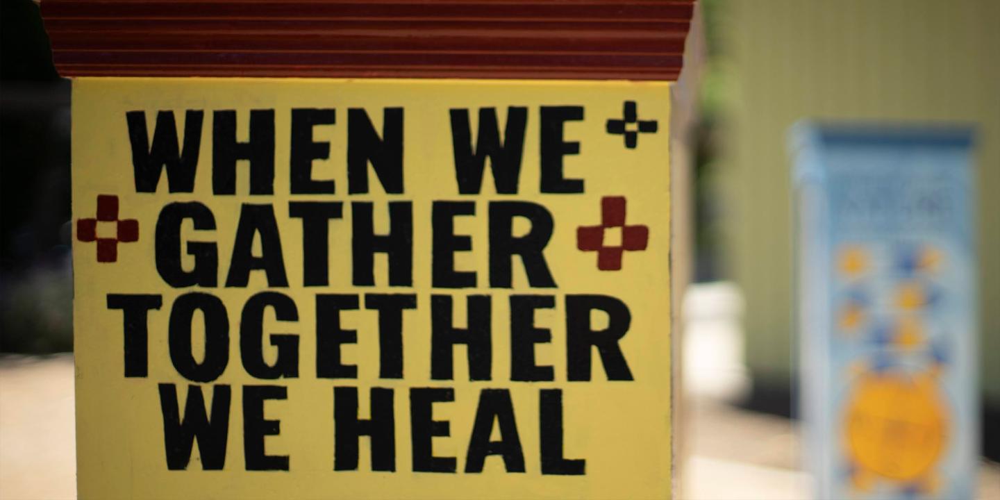 Photo of an outdoor artwork with the words, When We Gather Together We Heal, stamped in black letters on a yellow box.