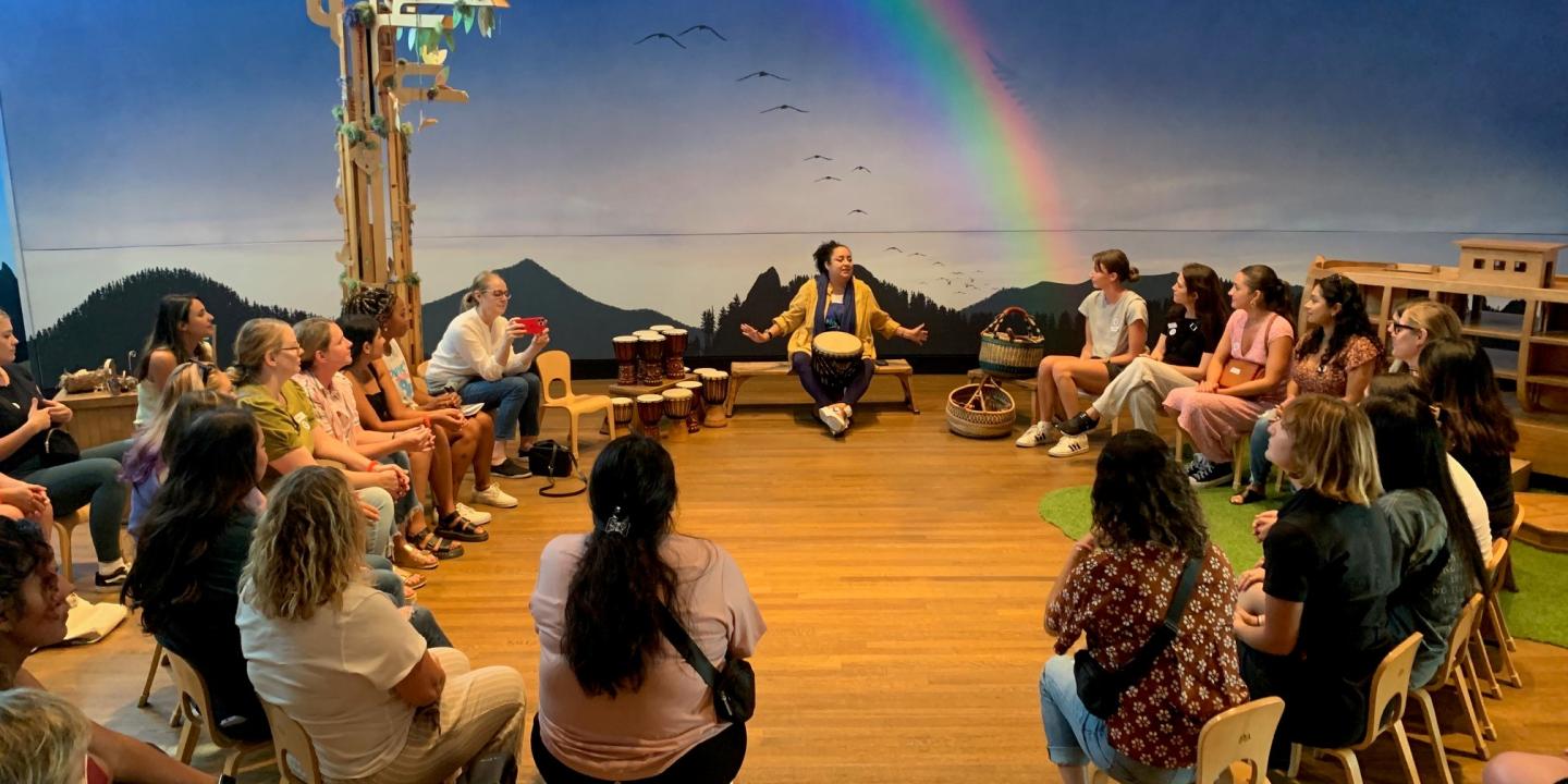 A group of people sitting on chairs in a circle. There is a wood floor. The blue wall in the back of the room has a rainbow painted on it.