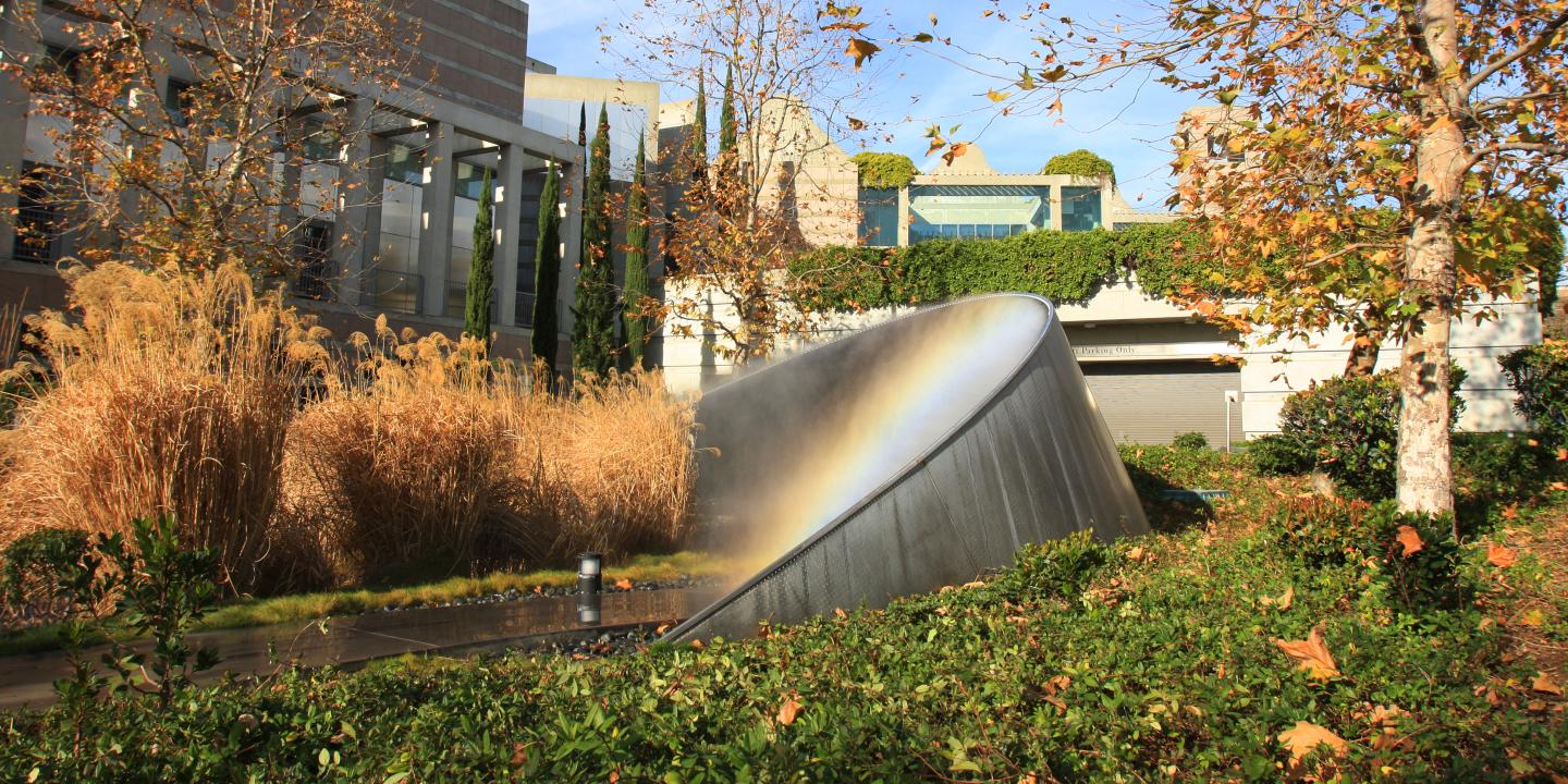 A metal sculpture that shaped in an arc emits mist and creates a rainbow. The sculpture sits in an green arroyo outside of a 3 story building.
