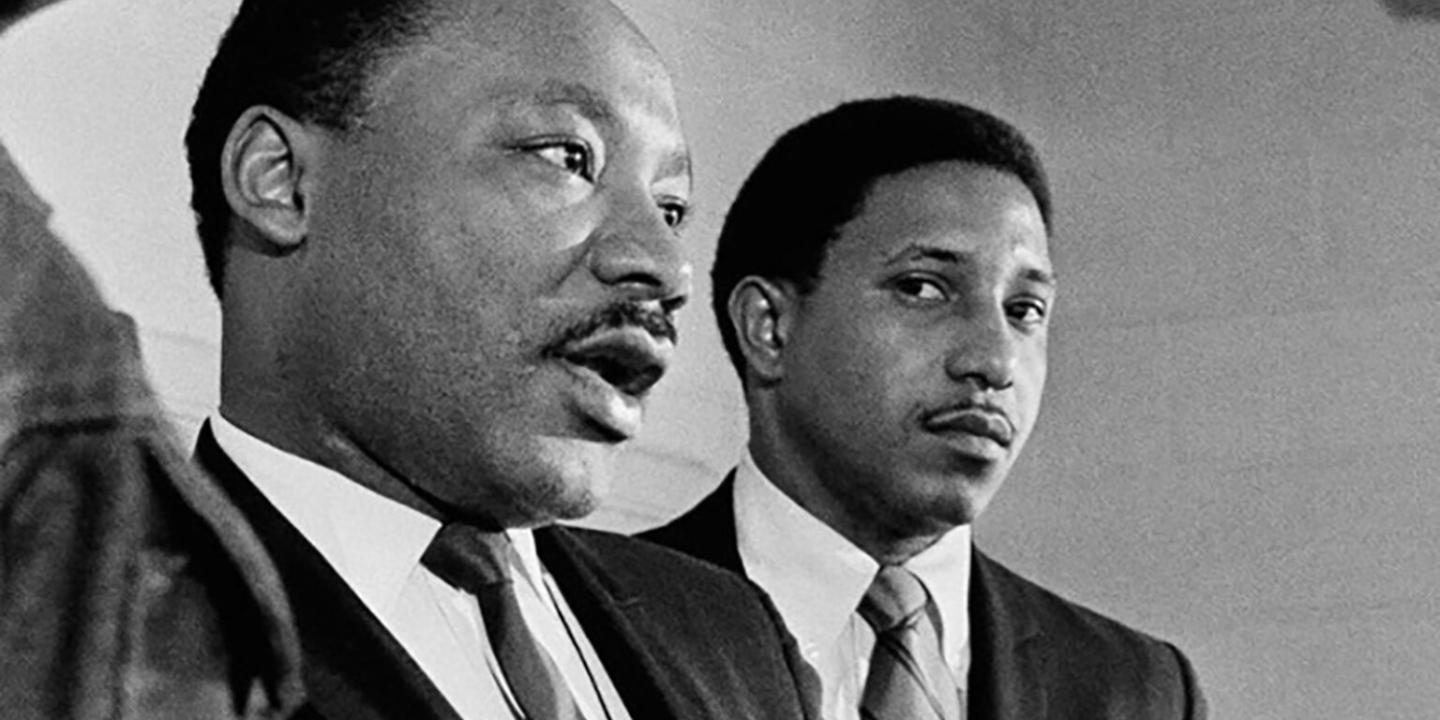 black-and-white photo of two African-American men dressed in suits and standing in front of microphones