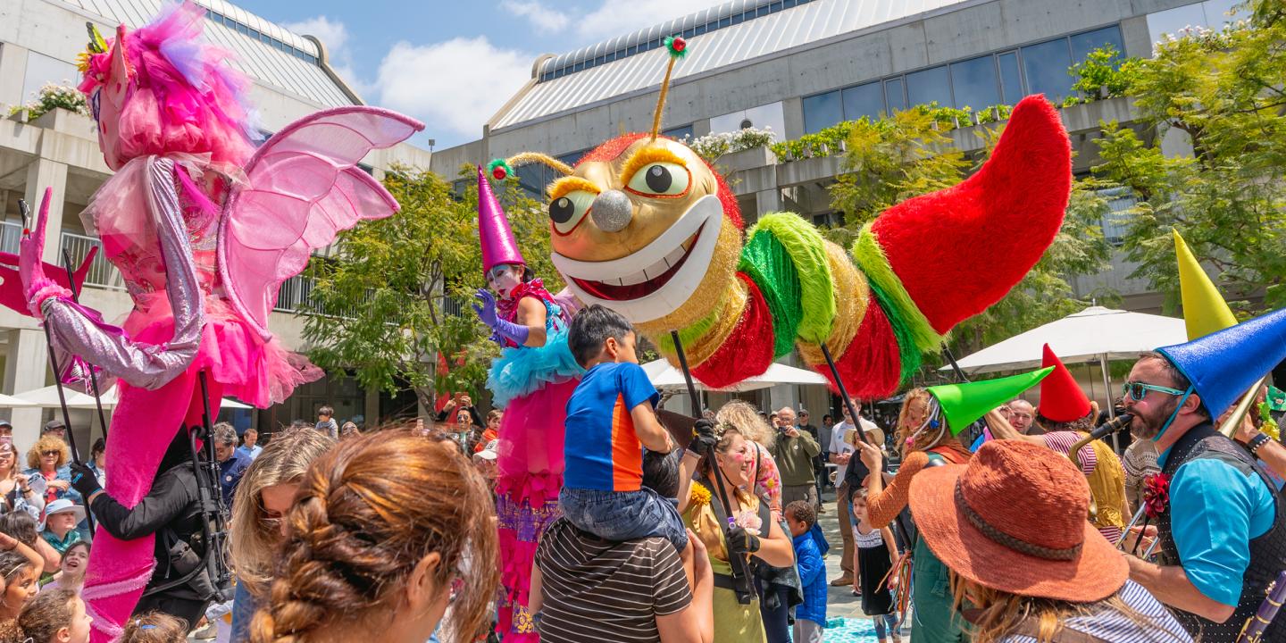 Children celebrating around a giant red and yellow caterpillar puppet being held by 3 adults with poles. 