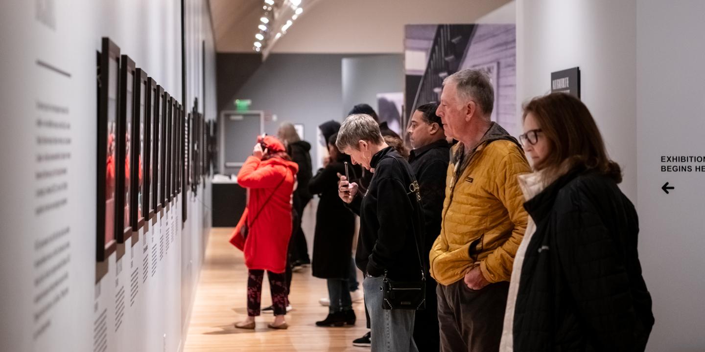 A large group of people standing in a line looking at a long wall of photos