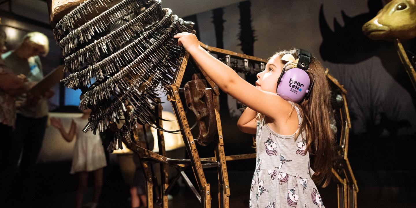 a young girl is wearing noise canceling headphones while interacting with a Noah's Ark deer made from keys and various materials.