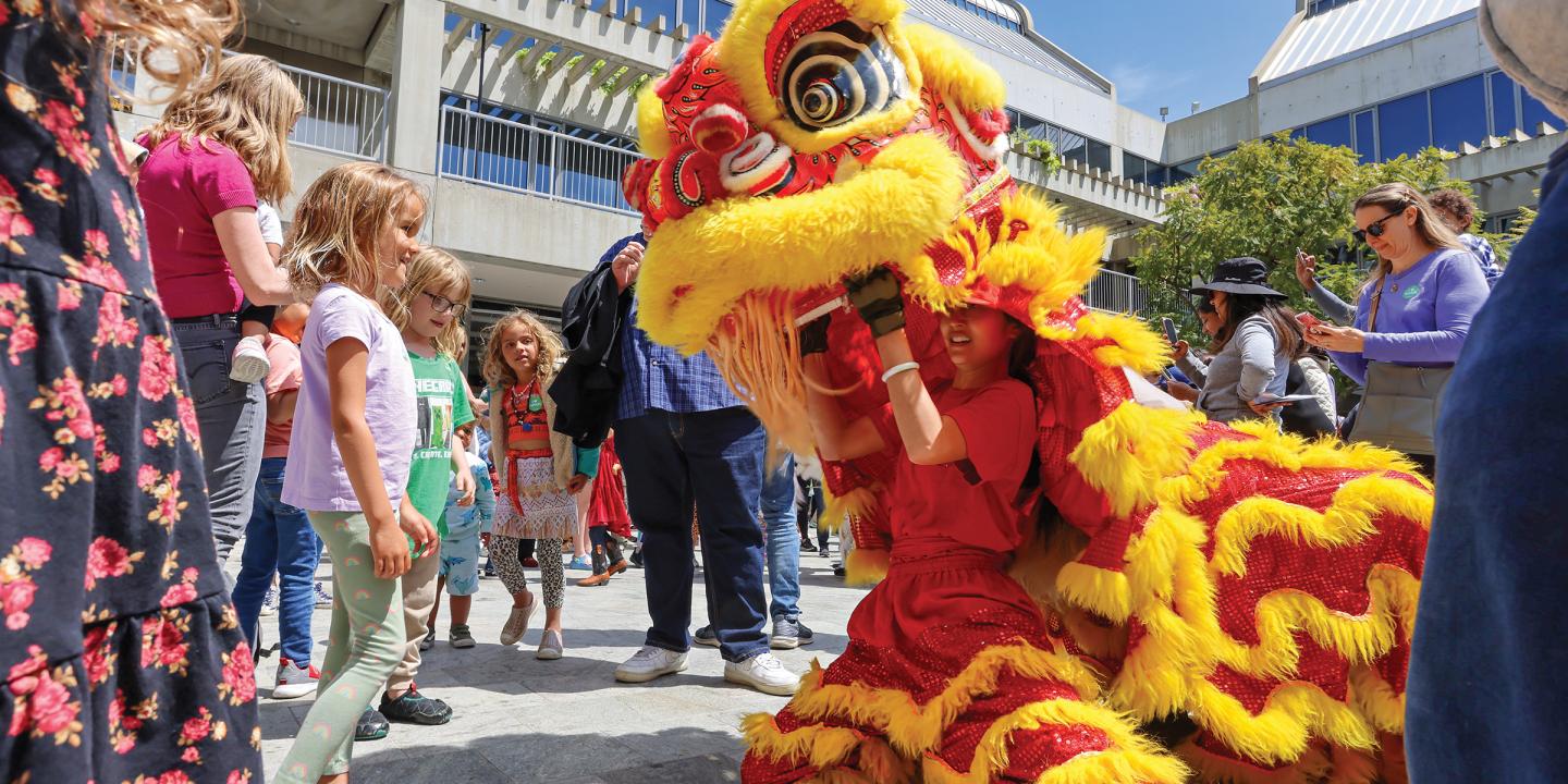 In a large, outdoor crowd, two young girls stand before a performer operating a large, red and yellow, dragon puppet