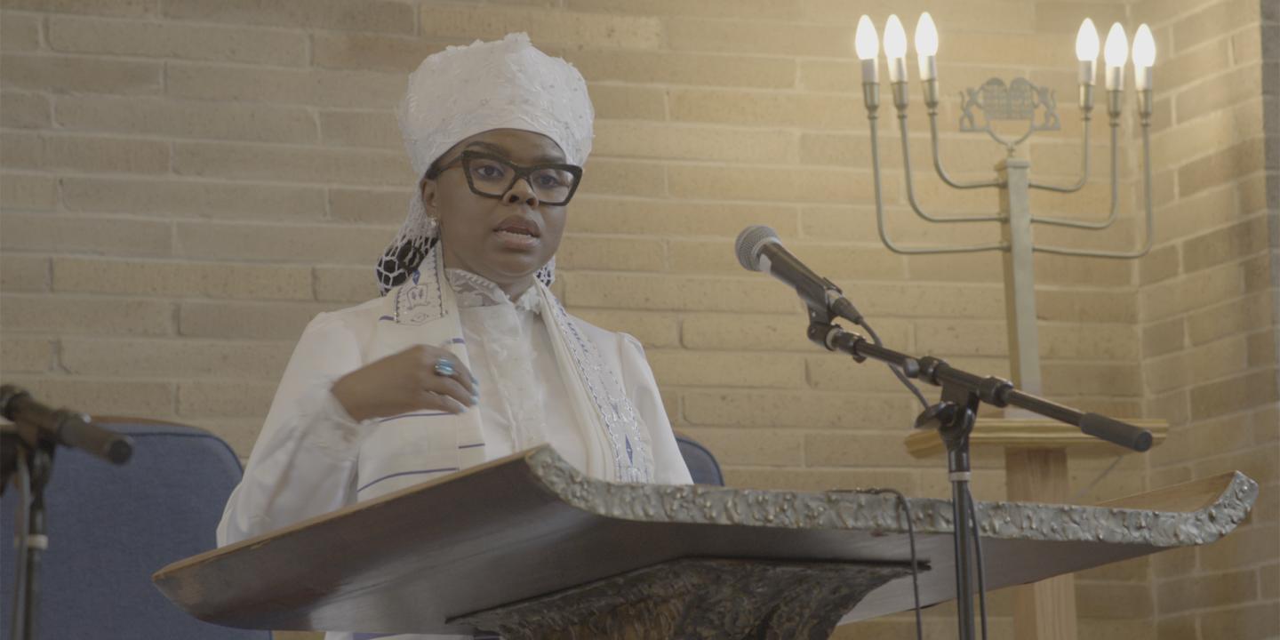 Rabbi Tamar Manasseh stands before a podium and microphone addressing a group of people out of frame. She is wearing all white attire at her ordination.
