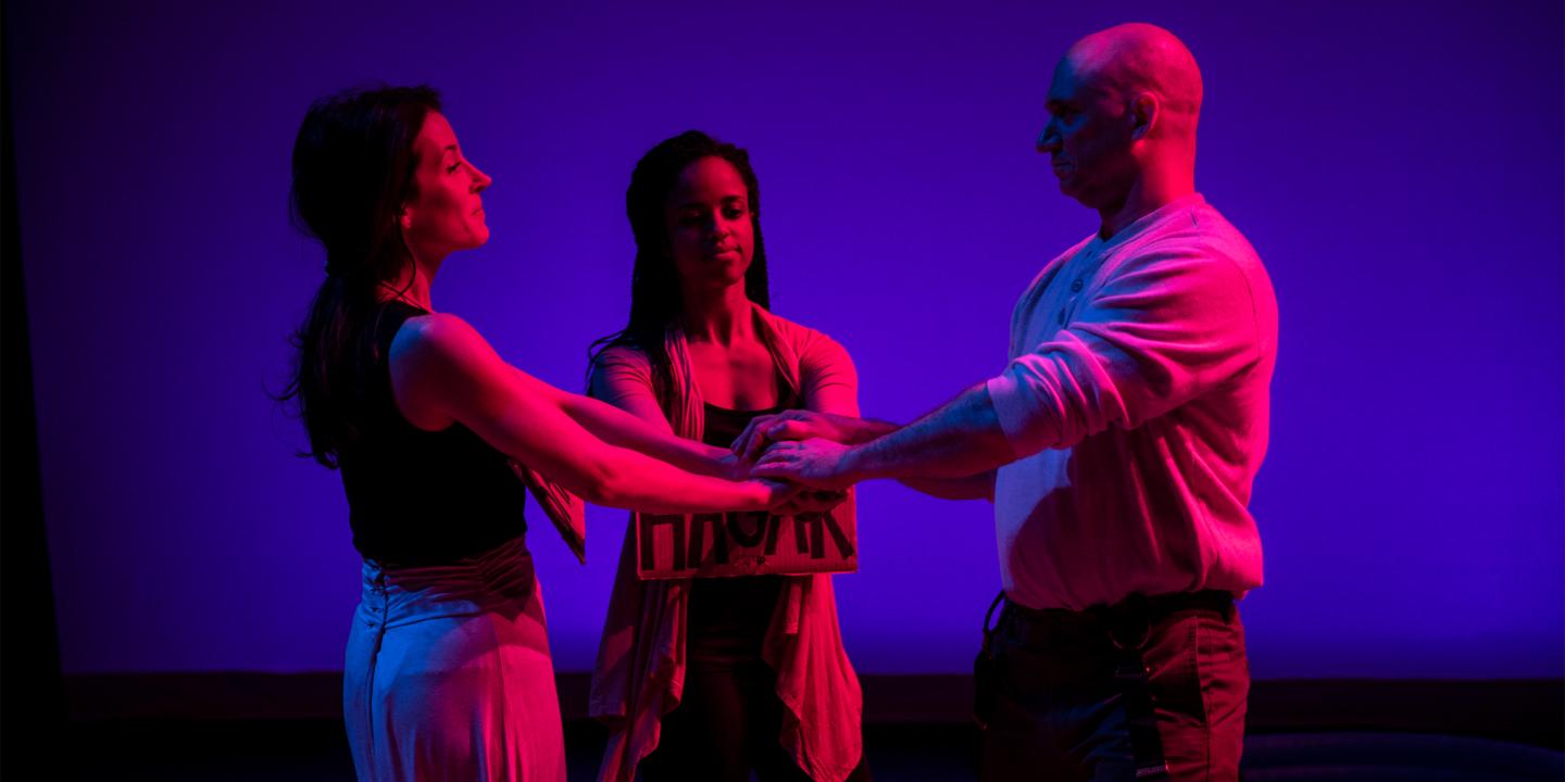 Three people stand on a stage facing each other and holding hand. They are lit with blue and red lights for a performance.