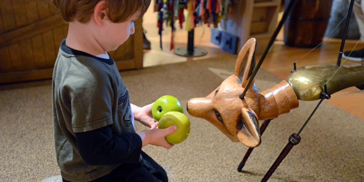 A young child gives two green toy apples to a puppet of a fox.