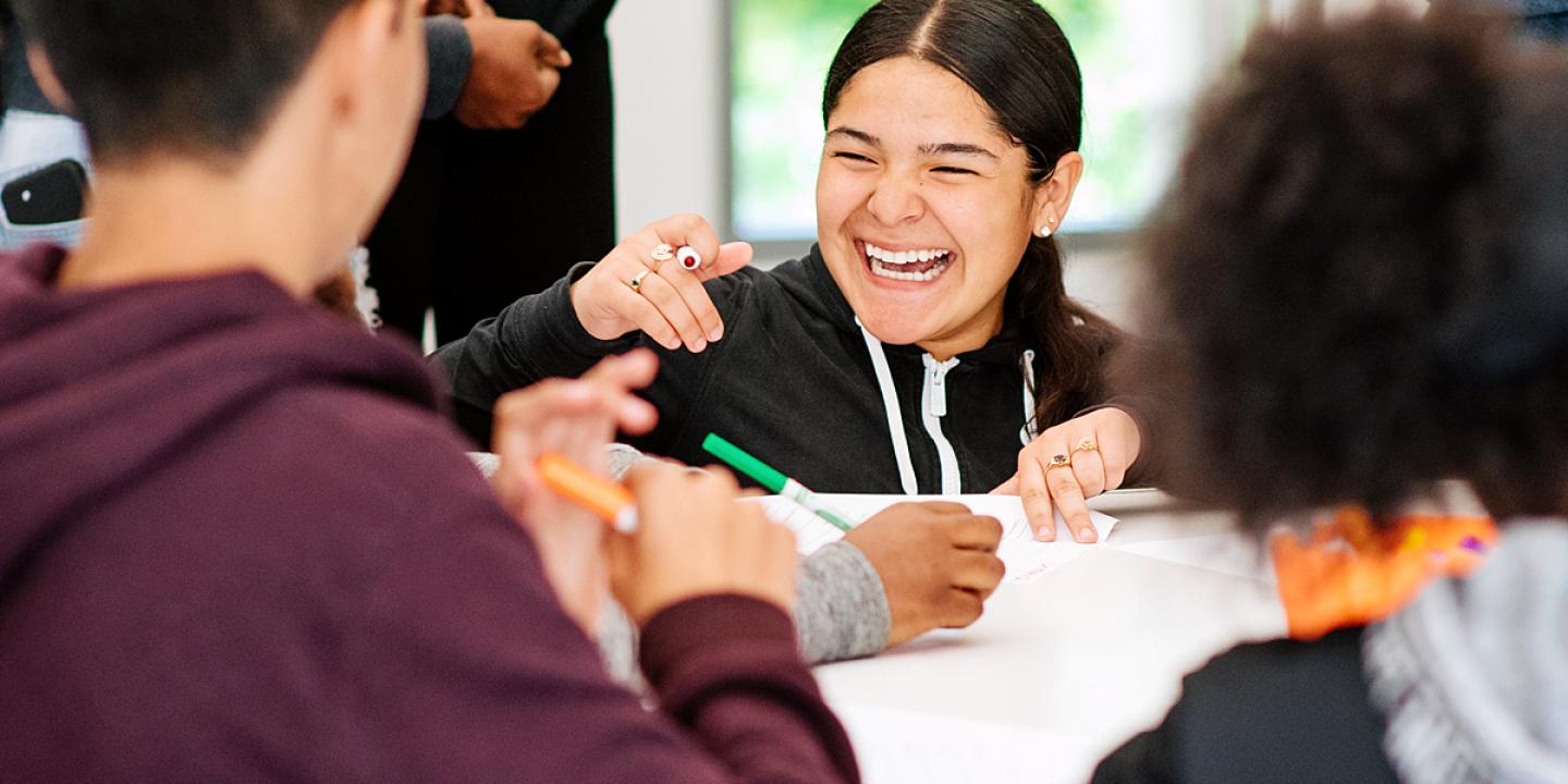 A teen smiling at a table while working with other teens.
