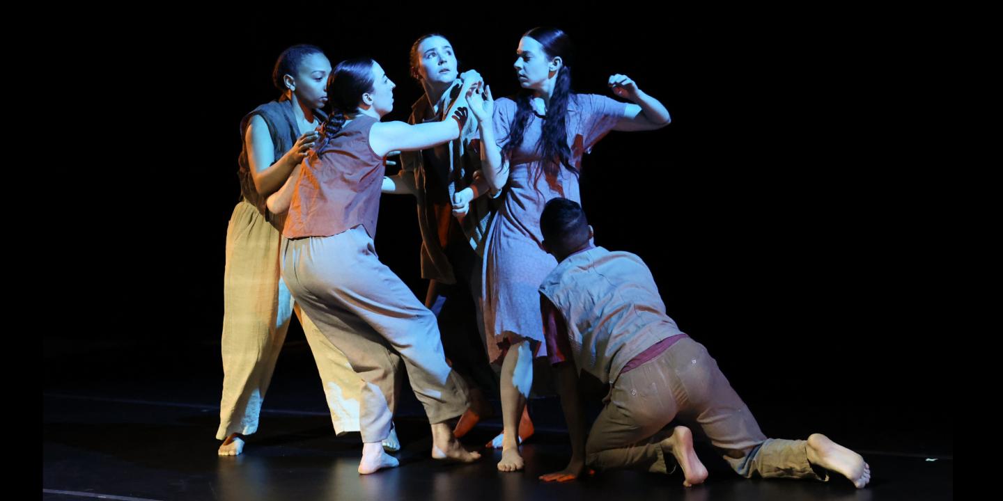A group of five dancers huddle together on a dark stage. One dancer faces outward, looking up.