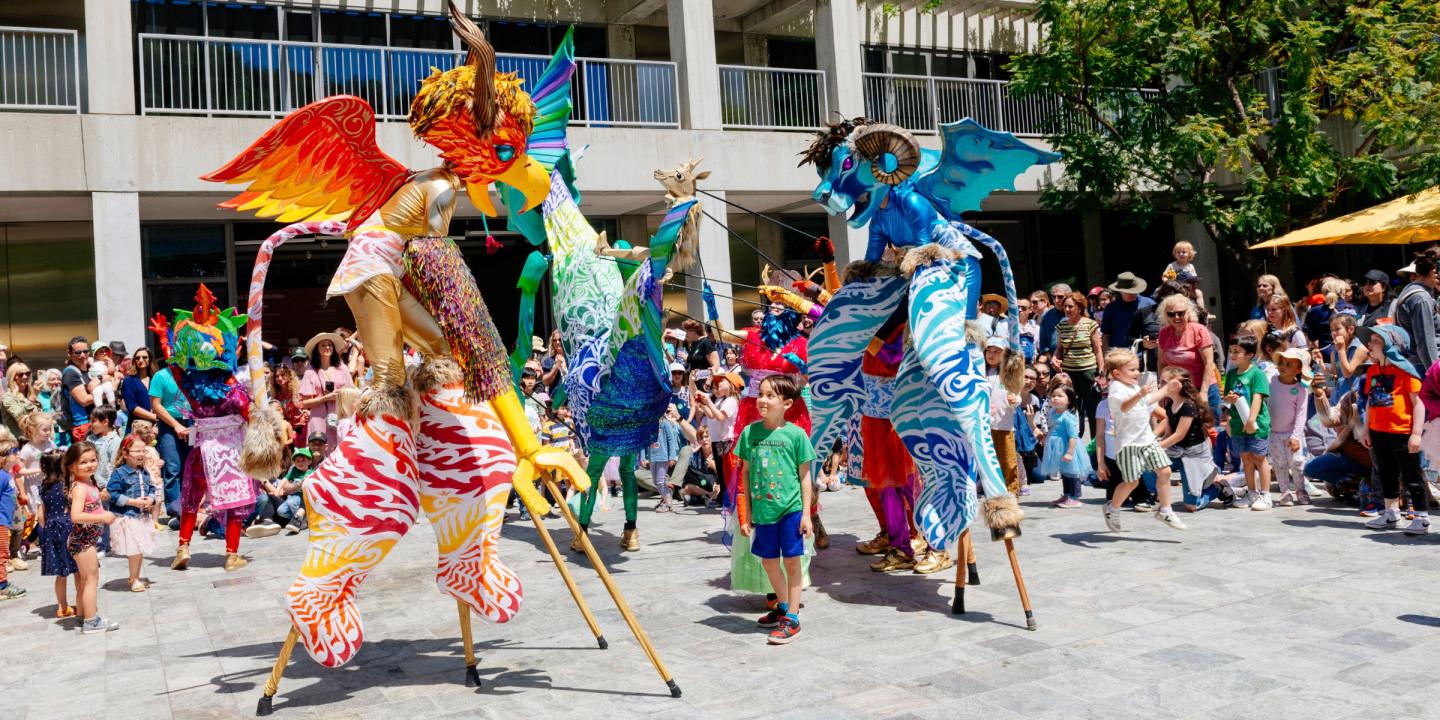 Performers on stilts in elaborate costumes move and dance in the middle of a large group of families in a Courtyard.