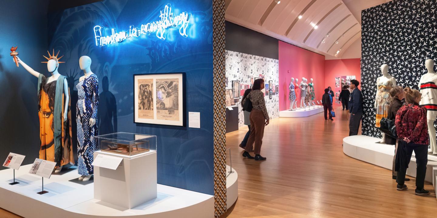 A gallery filled with people and clothing on mannequins. In the foreground are two mannequins dressed in different blue dresses, one holding a torch above its head like the Statue of Liberty.