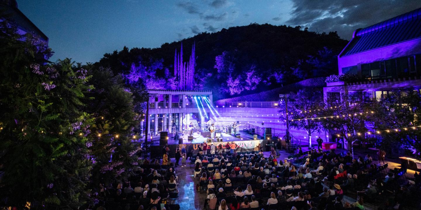 Photo of an outdoor concert at the Skirball. A large group of people are in the courtyard dancing to a band on stage. Lights are seen in the trees, strung across the courtyard, and from the stage.