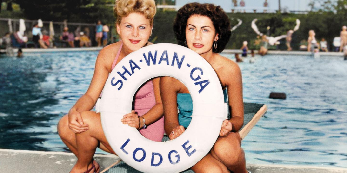 Two women pose in front of a swimming pool holding a life saver with the name Sha-Wan-Ga Lodge on it.