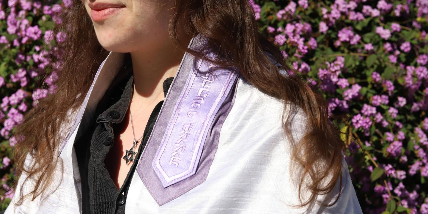 A woman wearing a purple tallit and silver Star of David necklace standing outside in front of a bush of purple flowers.