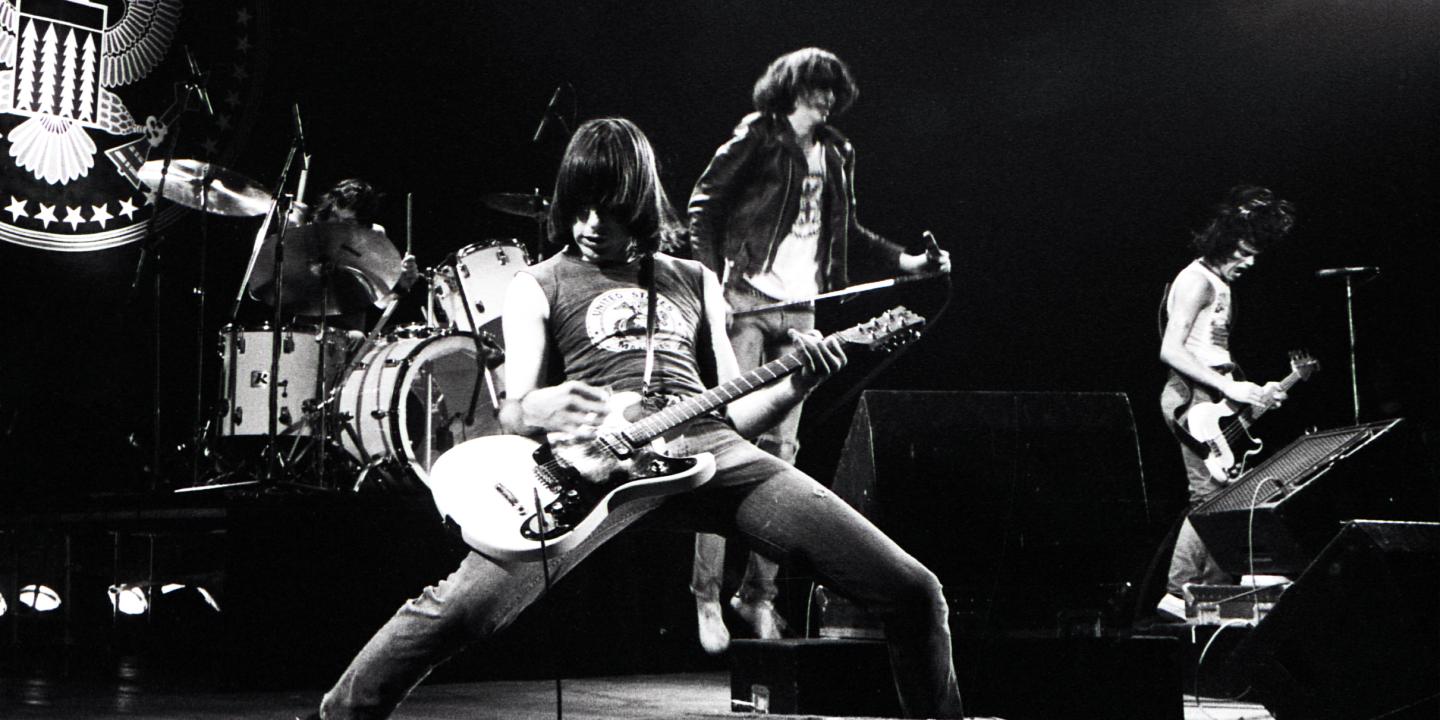 A black and white photo of a guitar player up front from his band on a stage playing.