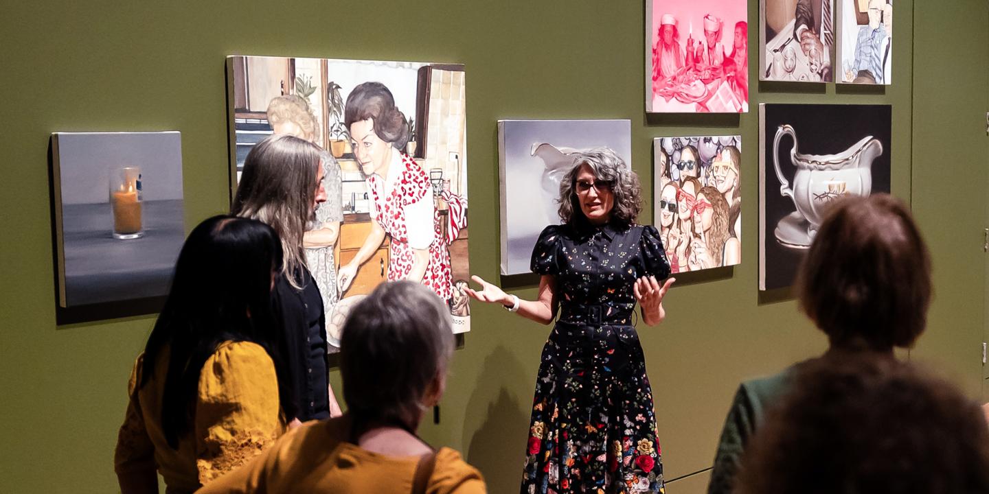A woman artists talks to a group of people in front of her painting hung in a gallery.