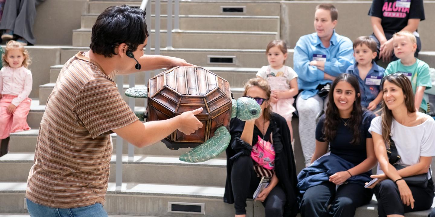 A man performs with a turtle puppet in front of families in an outdoor theater.