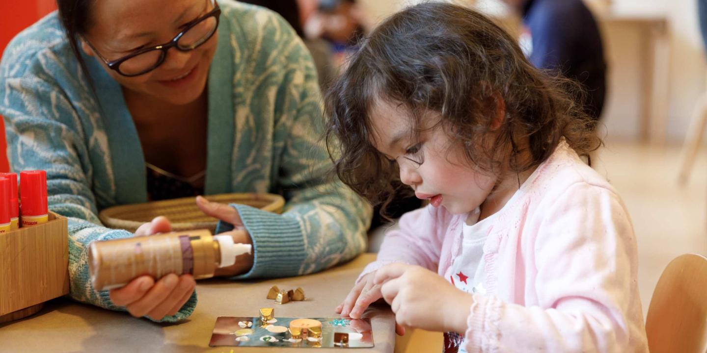 An adult helps a child on their craft project by holding the glue for them.