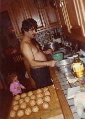 a shirtless man in a kitchen preparing food with a small girl standing behind him