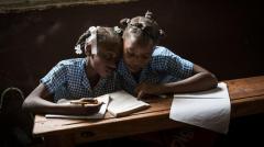 2 girls reading a book together and one girl has a pencil and is writing on a pad of paper