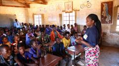 a teacher stands in front of a class of young boys and girls seated at desks