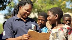 a woman holds a book next to 3 young students 