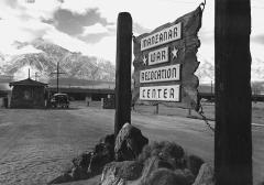 wooden sign that reads 'Manzanar War Relocation Center' with guard gate and car in the background