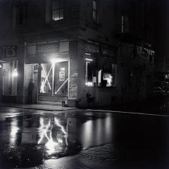 Black and white photo of shadowy man standing outside the doorway of a store at night